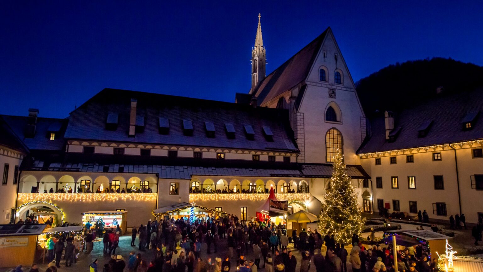 Adventmarkt im Mostviertel: beleuchtete Marktstände im Hof der Kartause Gaming mit Christbaum und Besuchern zur blauen Stunde